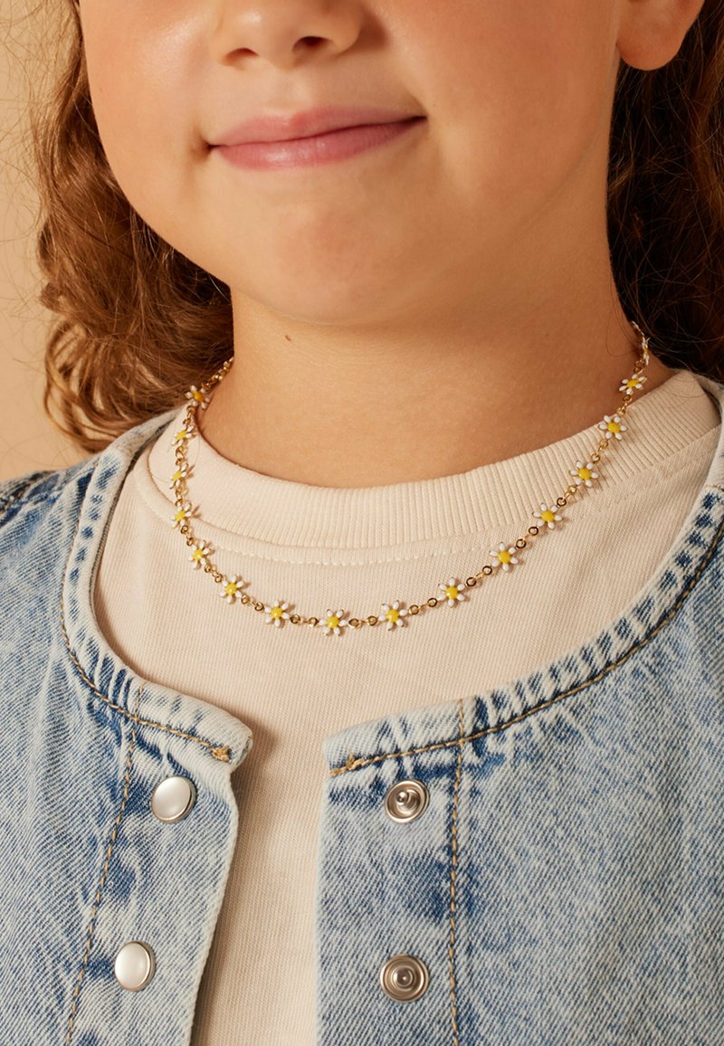 Gouden bloemenketting met kleine witte en gele madeliefjes, gedragen over een crèmekleurige sweatshirt onder een lichtblauwe spijkerjack.