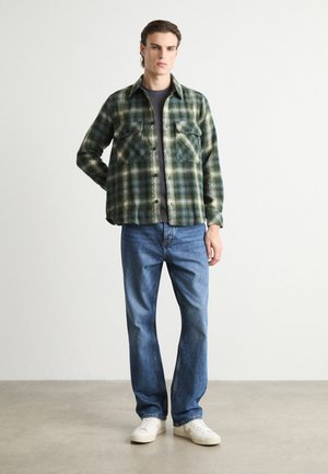 Green and white plaid shirt with button closure and two chest pockets, paired with light blue jeans and white sneakers. Smooth fabric texture.