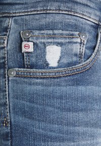 Wycieniowane niebieskie dżinsy z wystrzępioną tylną kieszenią z małym szwem i srebrnymi metalowymi elementami, w tym guzikiem i nitami.