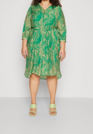 Frau trägt ein grünes knielanges Kleid mit beigen Blattmustern und hellgrünen Sandalen mit offenen Zehen, steht vor einem schlichten Hintergrund.