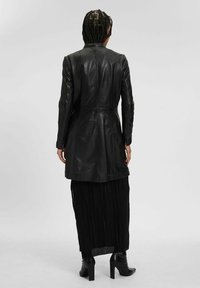 Cappotto in pelle nera con vita aderente, design a metà lunghezza e texture liscia. Indossato sopra una gonna nera plissettata, abbinato a stivali con tacco alto.