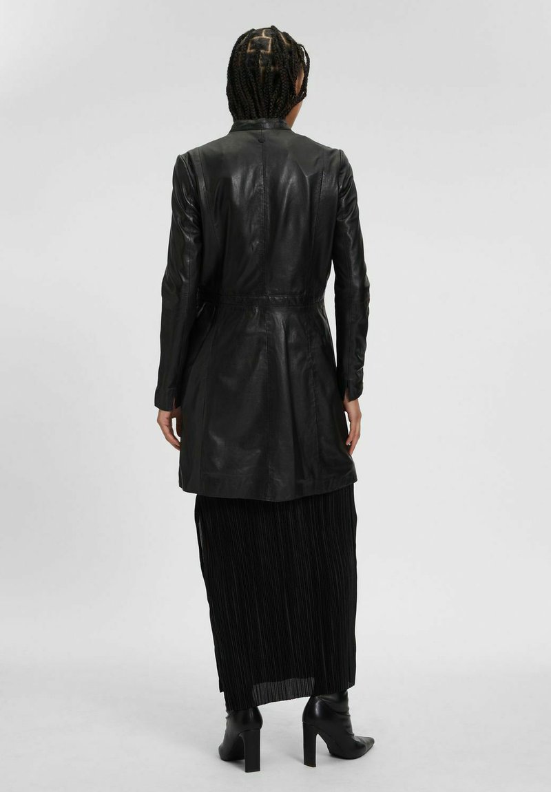 Cappotto in pelle nera con vita aderente, design a metà lunghezza e texture liscia. Indossato sopra una gonna nera plissettata, abbinato a stivali con tacco alto.