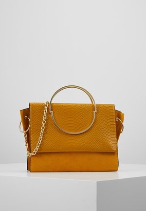 Sac à main jaune moutarde avec rabat texturé, grande anse circulaire dorée et bandoulière chaîne dorée attachée sur une surface blanche sur fond gris.