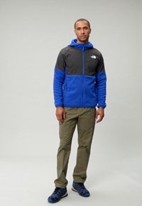 Fleece striukė su pilkais ir mėlynais dėmiais, užtrauktuku priekyje, su kapuce ir logotipu. Alyvuogių žalios spalvos kelnės ir juodo-mėlyno dizaino sportbačiai užbaigia aprangą.