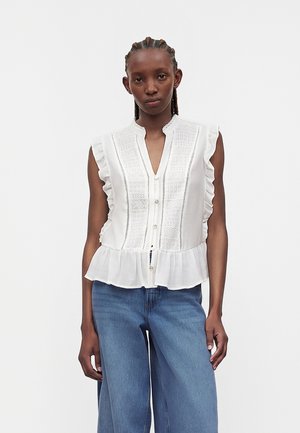 Woman with braided hair wearing a sleeveless white ruffled blouse with embroidery and blue wide-leg jeans, standing against a plain background.