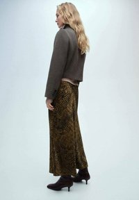 Braune, taillierte Jacke über einem hellrosa Oberteil, kombiniert mit einem langen, gemusterten Rock in dunklem Olivgrün und Braun. Mit braunen Knöchelstiefeln gestylt.