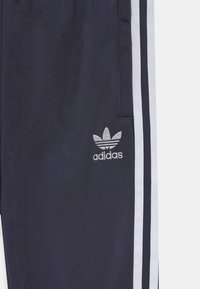 Námořnické atletické kalhoty s bílými bočními pruhy a vyšitým logem Adidas. Vyrobeny z hladké, odolné tkaniny s tapered střihem a kapsami na zip.