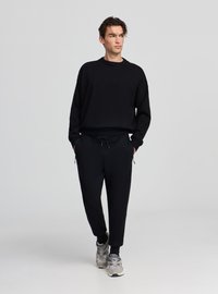 Completo felpa nera e pantaloni jogger, tessuto morbido, vestibilità rilassata, polsini e vita elasticizzati, dotato di tasche con cerniera, abbinato a scarpe da ginnastica grigie.