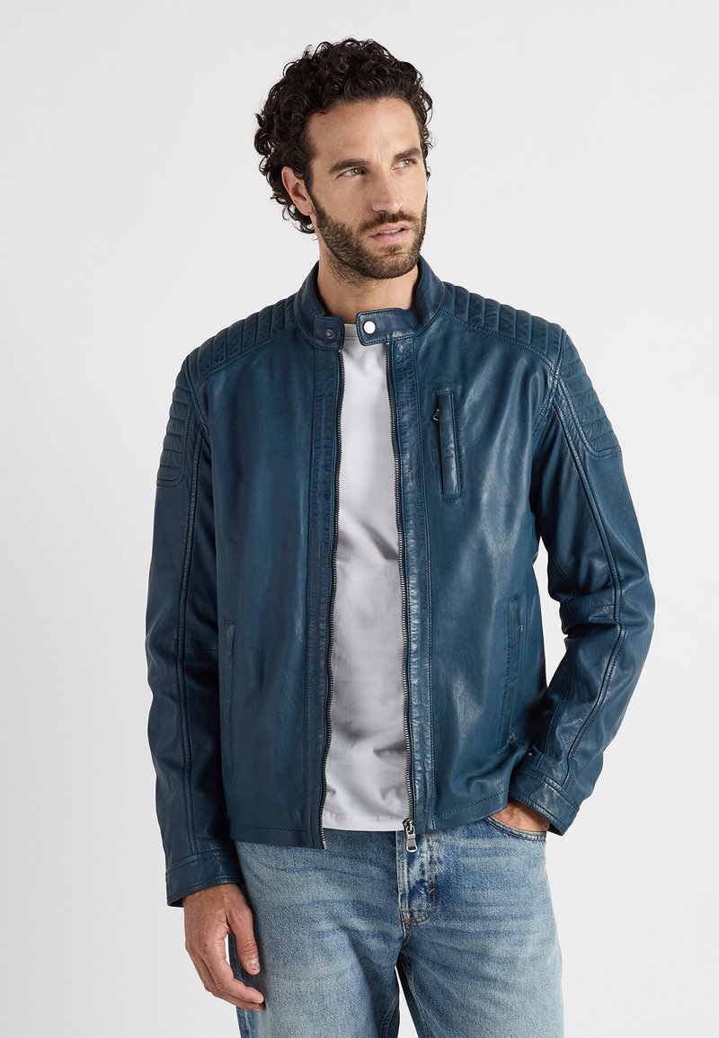 Blauwe leren jas met een opstaande kraag, gestikte schouderdetails, een rits aan de voorkant en een borstzak, gedragen over een wit t-shirt.