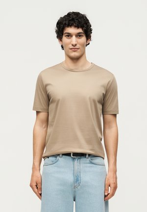 Jeune homme aux cheveux courts, bouclés et foncés, portant un t-shirt beige à manches courtes et un jean bleu clair, debout devant un fond uni clair.