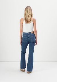 Blond kvinna som står vänd bort, iklädd en vit ärmlös topp, blå högmidjade jeans och beige skor mot en enkel vit bakgrund.