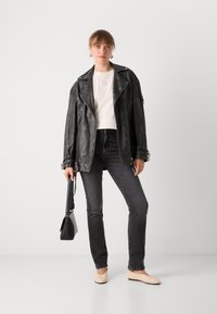 Zwarte leren motorjack, witte getailleerde top, donkere spijkerbroek, roomkleurige flats en een kleine zwarte handtas; model staat tegen een plain witte achtergrond.
