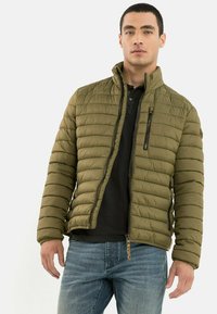 Olivengrøn quilted pufferjakke med høj krave, frontlynlås og sidelommer. Kombineret med en sort skjorte og blå jeans.
