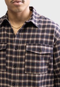 Cămașă din flanel în carouri de culoare albastru închis, maro și galben. Dispune de două buzunare frontale cu nasturi și un guler clasic. Țesătură texturată.