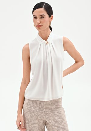 Donna che indossa una camicetta bianca senza maniche con pieghe e spilla di perle, abbinata a pantaloni beige con tessuto strutturato, che posa davanti a uno sfondo semplice.