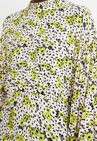 Robe à motifs avec un fond crème orné de motifs floraux noirs et verts vifs, tissu léger, col montant et taille froncée.