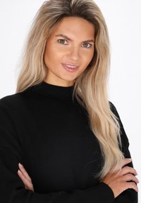 Pulover negru cu guler înalt, textură moale. Păr lung, ondulat, blond, expresie zâmbitoare, brațele încrucișate, fundal neutru.