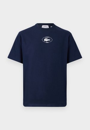 Mand og kvinde står op, begge iført marineblå t-shirts med grafik; manden i lyseblå jeans og hvide sneakers, kvinden i beige shorts og hvide sneakers.