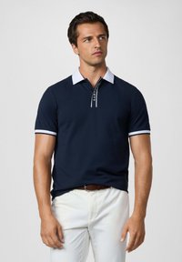 Námornícky polo tričko s bielym golierom a pruhovanými manžetami. Obsahuje polovičný zips a logo na léze. Hladká textúra tkaniny.