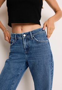 Højtaljede blå denim jeans med en falmet vask, fem-lomme design og en rå kant. Stoffet ser robust og struktureret ud.