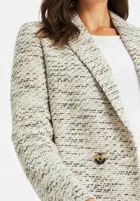 Kobieta w teksturowanej beżowo-czarnej marynarce z tkaniny Tweed, z pojedynczym guzikem w kolorze muszli żółwia, nosząca białą bluzkę.