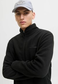 Giacca in pile nera con colletto alto e frontale con zip. Presenta una piccola tasca orizzontale e una texture morbida. Abbinata a un cappello grigio chiaro.