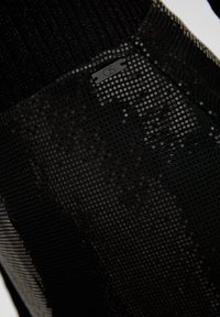 Schwarzer Netzstoff mit einer strukturierten Oberfläche, der runde Aussparungen und ein kleines QS-Etikett aufweist. Das Gesamtdesign ist schlank und modern.