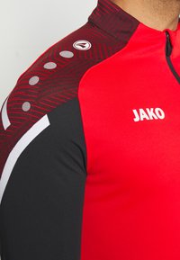 Rood en zwart sporttop met een hoge kraag, ritsluiting en een gestructureerd patroon met ronde accenten op de schouders. JAKO-logo in het wit.