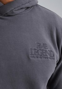 Sudadera gris hecha de tejido suave con un bolsillo tipo canguro. Presenta un texto de logotipo en relieve que dice "RVE LEGEND" en el pecho, con un corte relajado.
