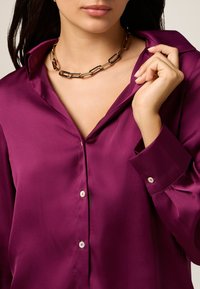 Paarse satijnen overhemd met knopen en kraag, met een relaxte pasvorm en zichtbare knoopdetails. Draagt een chunky gouden ketting als accessoire.