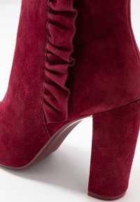 Stivaletto a tacco alto in suede bordeaux con dettaglio arricciato lungo il lato e tacco quadrato, caratterizzato da una texture liscia e opaca.