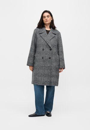 Femme debout portant un manteau gris à carreaux croisé, un jean bleu pattes larges et des chaussures plates noires sur un fond uni.