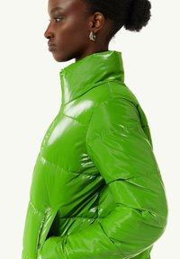 Glanzend, felgroen pufferjack met hoge kraag, doorgestikte uitstraling en zijzakken. De stoftextuur lijkt glad en glanzend.