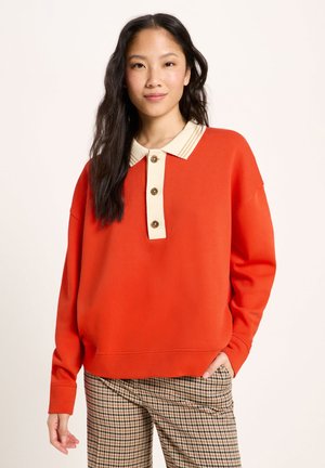 Frau mit langen dunklen Haaren, die ein orangefarbenes langärmliges Poloshirt und beige karierte Hosen trägt und mit einer Hand in der Tasche posiert.
