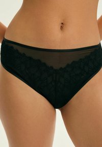 Czarny koronkowy stanik w stylu bikini z półprzezroczystą siateczką i haftem kwiatowym noszony na opalonej talii i biodrach na jednolitym tle.