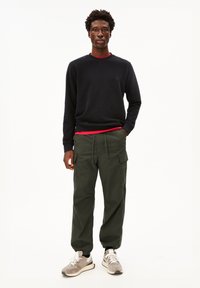 Felpa nera con scollatura a girocollo, indossata sopra una maglietta rossa. Pantaloni cargo verde oliva con tasche laterali, abbinati a sneakers grigie.