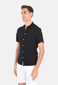 Camicia a maniche corte con bottoni in tessuto blu navy uniforme, con colletto alla cinese. Include bottoni bianchi e una trama leggera. Abbinata a pantaloni corti bianchi.