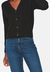 Black knit cardigan with a V-neck, featuring six dark buttons and long sleeves, paired with blue denim jeans. Smooth texture and fitted shape.