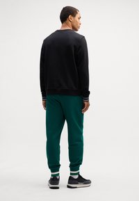 Felpa nera con scollo rotondo, abbinata a pantaloni della tuta verdi con polsini a righe. Completato con scarpe da ginnastica nere.