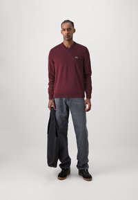 Burgundy színű V nyakú pulóver hosszú ujjakkal, kék farmernadrág felett viselve. A model fekete táskát hord, fekete cipőket visel világosbarna talppal.
