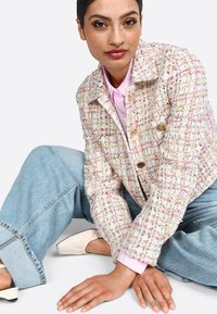 Giacca in tweed testurizzato color crema con macchie multicolori, design doppiopetto, abbinata a jeans baggy azzurri chiari e una camicia rosa.