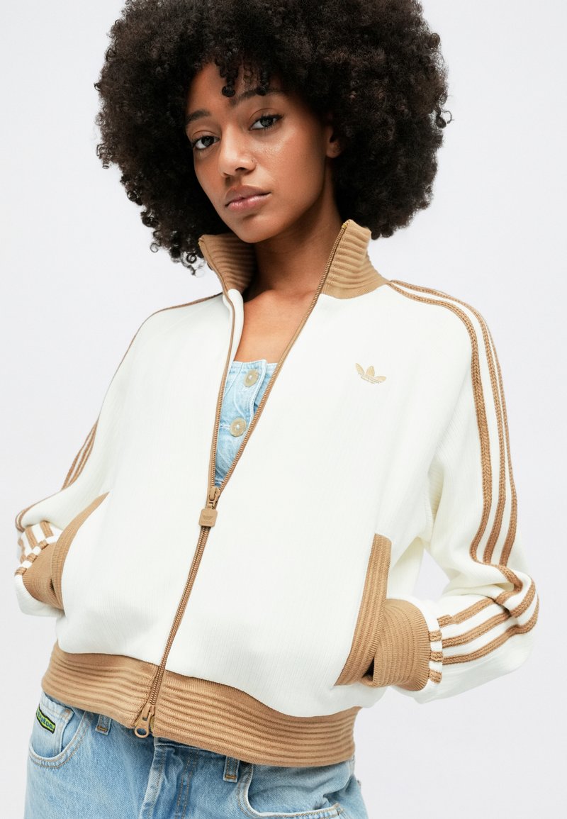 Veste zipée blanche avec des accents marron, poignets et ourlet côtelés, dotée de trois rayures sur les manches et d'un logo brodé sur la poitrine.