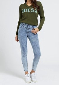 Femme portant un pull GUESS vert olive, un jean skinny taille haute bleu clair et des baskets plateformes blanches, se tenant devant un fond uni.