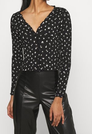 Femme portant un chemisier noir à manches longues boutonné avec de petits motifs floraux blancs et un pantalon en cuir noir taille haute.