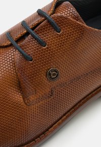 Scarpa in pelle marrone con texture e motivo esagonale, dotata di lacci neri e un piccolo logo bronzeo sul lato.