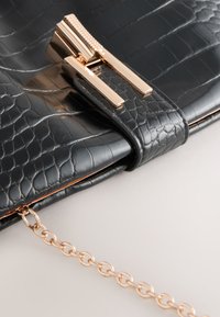 Crna torbica teksture krokodilske kože sa zlatnim kopčanjem i nježnim zlatnim lancem na svijetloj pozadini.