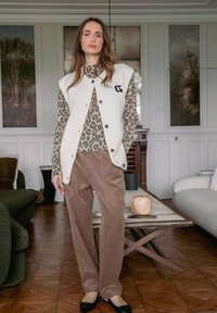 Mujer vistiendo una blusa de manga larga con estampado de leopardo, un chaleco blanco sin mangas con botones, pantalones de pana marrones y zapatos planos negros en una sala de estar.