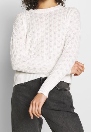 Kvinde iført en hvid kabelstrikket sweater med hjerte-mønstre og sorte højtaljede jeans, stående med armene krydset.