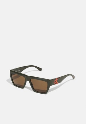 Rechteckige Sonnenbrille mit braunen Gläsern, olivgrünem Kunststoffrahmen und rotem CK-Logo an der Schläfe. Glatte Textur und modernes Design.