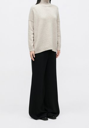 Personne portant un pull à col roulé beige avec des manches longues et un pantalon noir large et ample, debout devant un fond blanc uni.